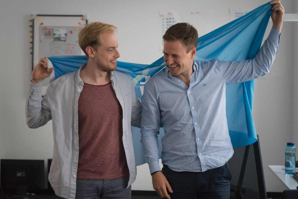 2 men looking at each other laughing while holding a joinmytrip blue flag.