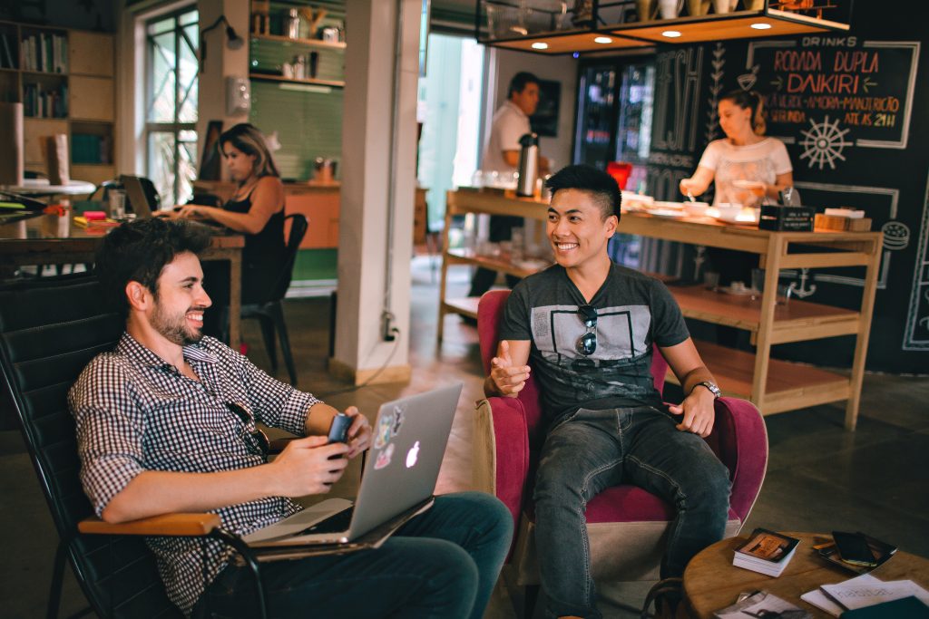 2 men sitting on a chair laughing and talking while three people in the background are cooking.