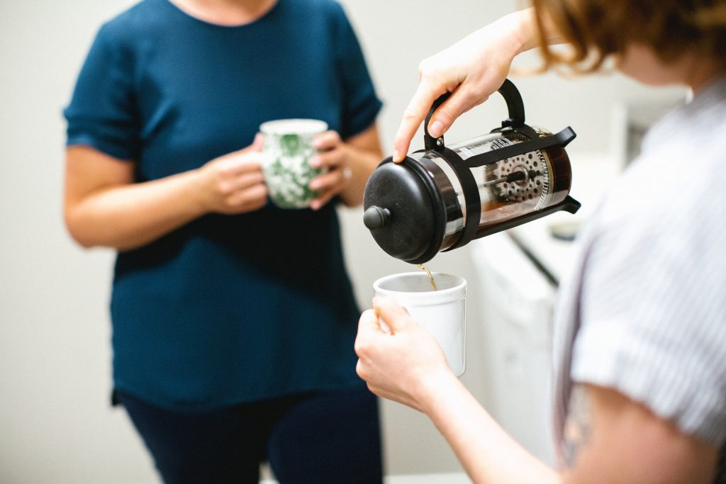 two woman standing in a kitchen with cups in their hands while one of them pours coffee.