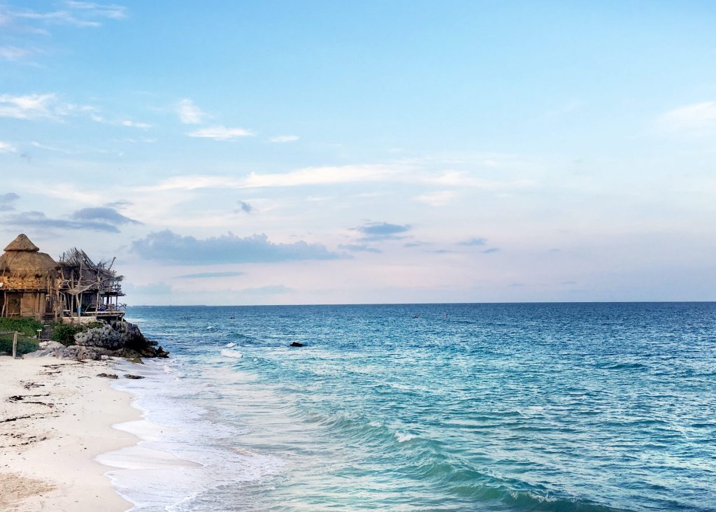 A beach in Tulum with beautiful blue water washing over it, blue skies and a Mayan temple