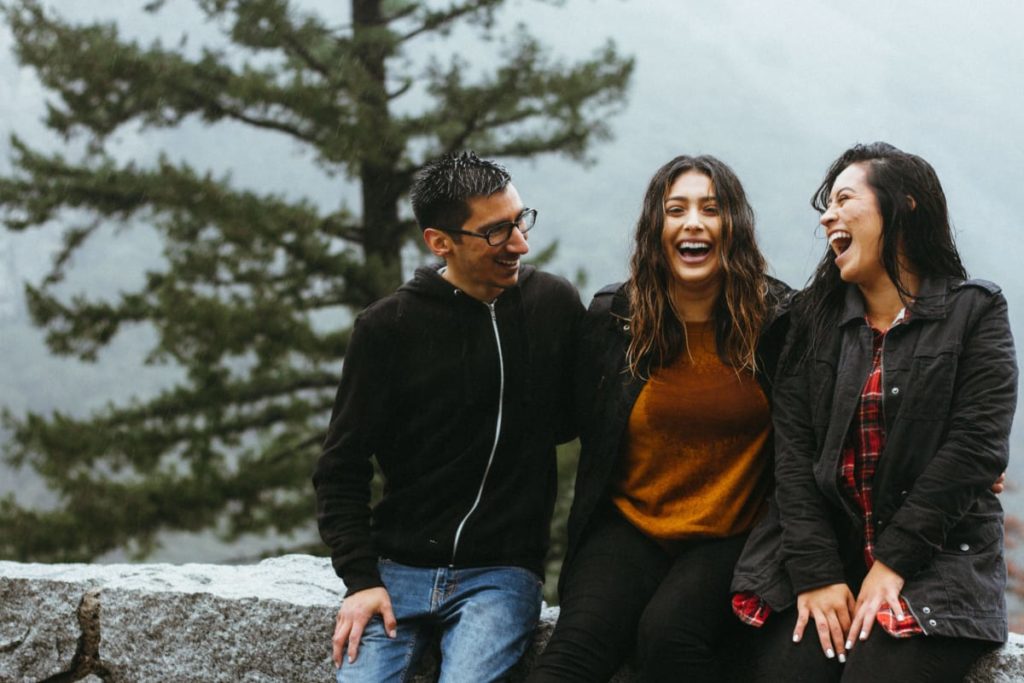 a man and two woman sitting on a stone wall laughing with a tree behind because they are traveling after corona