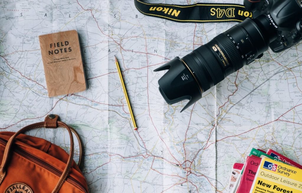 Orange backpack, a small brown notebook, travel magazines and a Nikon D4s camera lying on a street map 