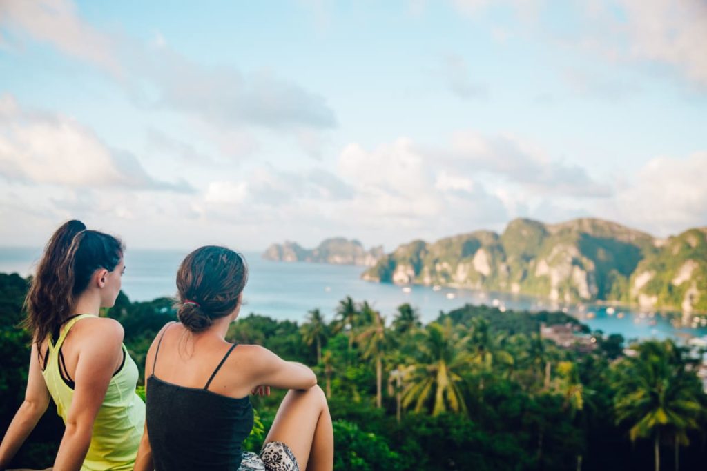 2 girls sitting together while looking at the view of the sea and mountains and forests