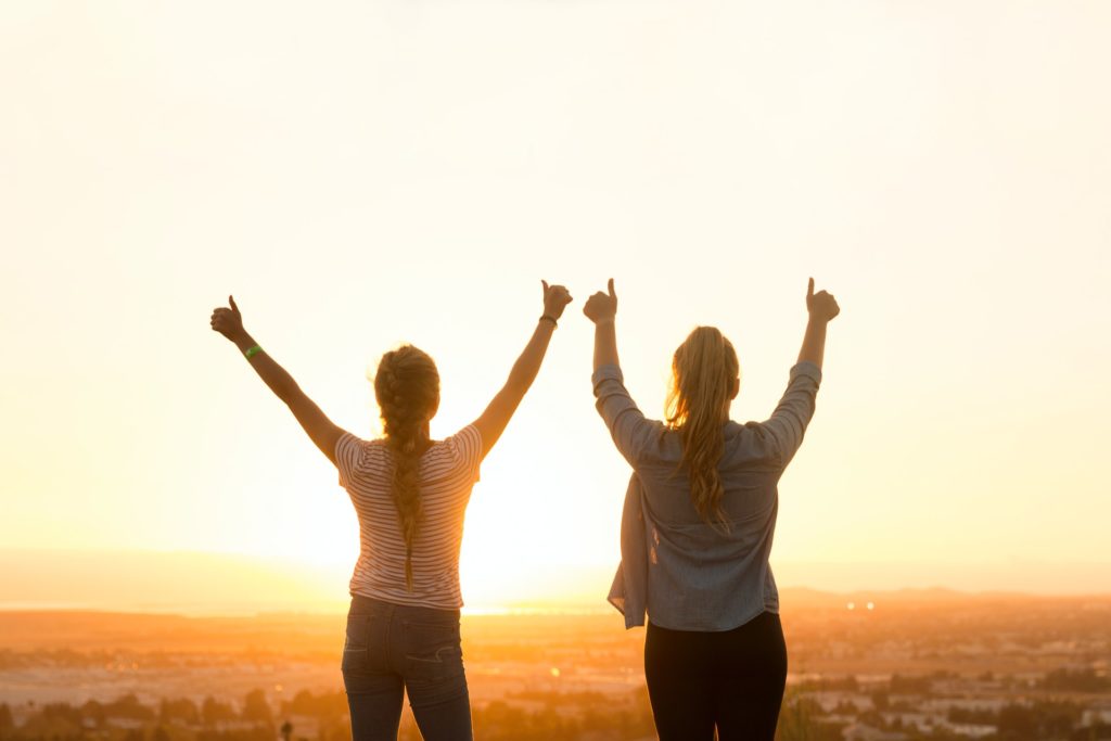 two women traveling and stood looking forward holding their thumbs up in front of a beautiful sunset 