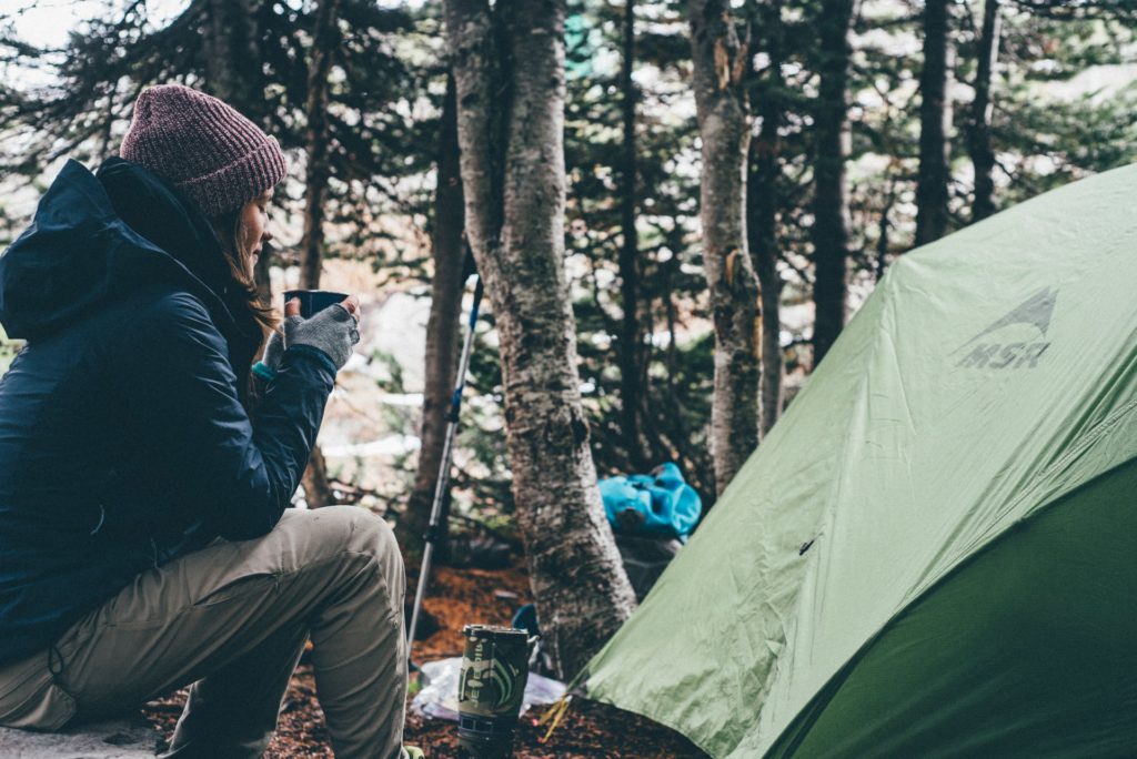 female, woman sat by tent holding a mug of tea, keeping warm while being surrounded by beautiful forests while camping on a hiking trip in germany