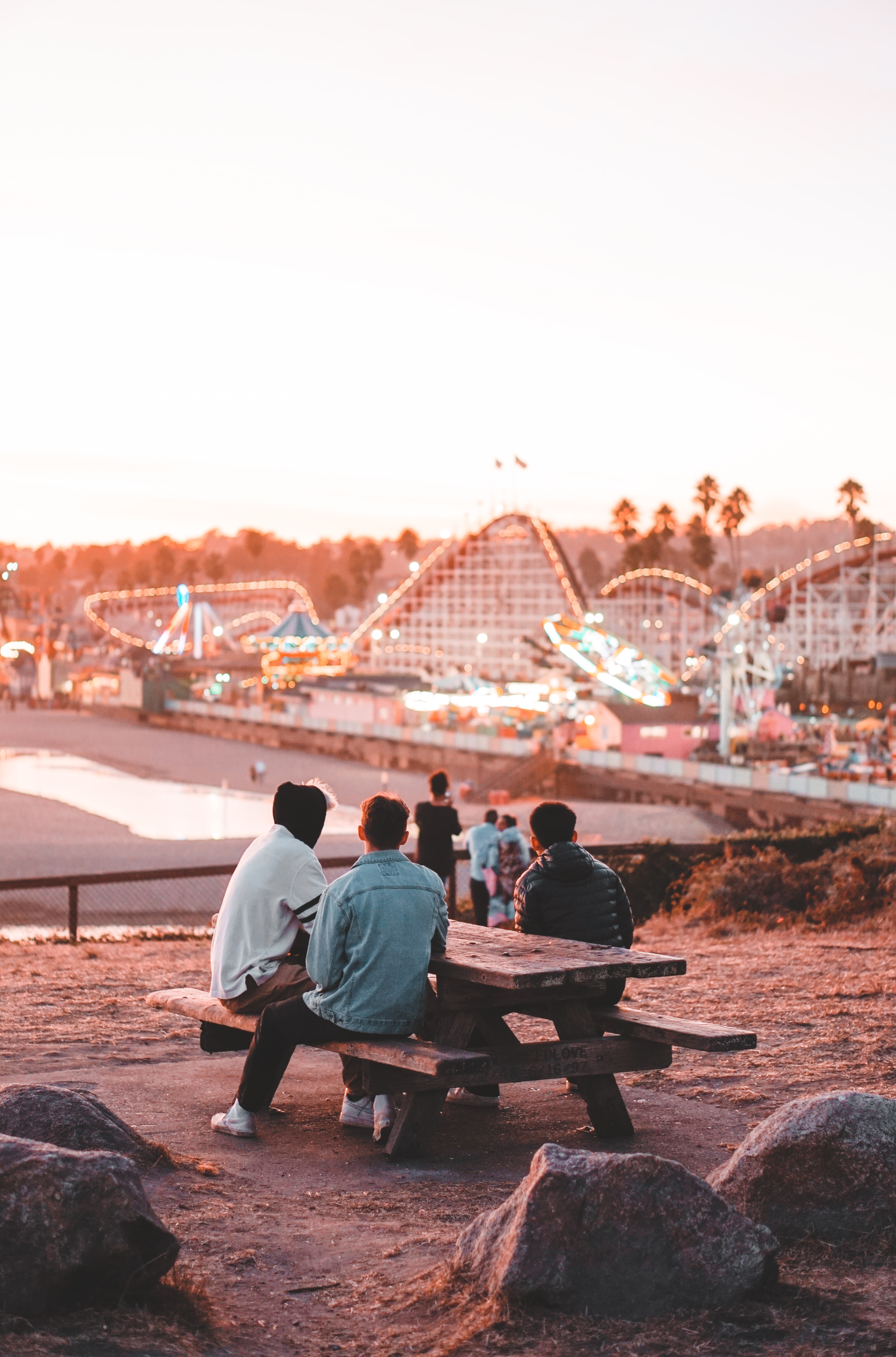 A group of friends is sitting on a bench in a theme park. In the background you can see roller coasters and other rides. 