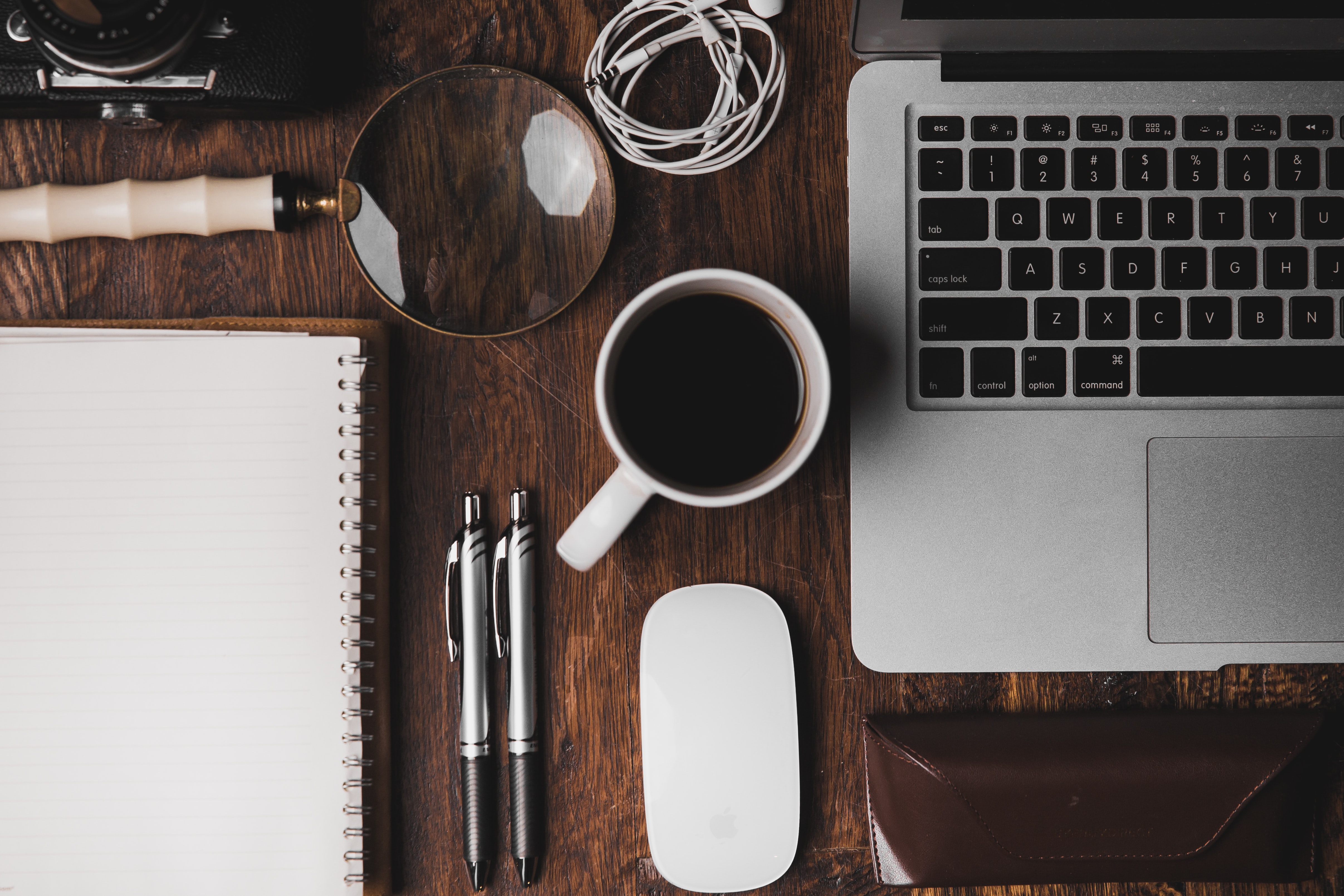 The top essentials for your coworking trip is a laptop, notebook, pens and coffee on top of a brown wooden table.